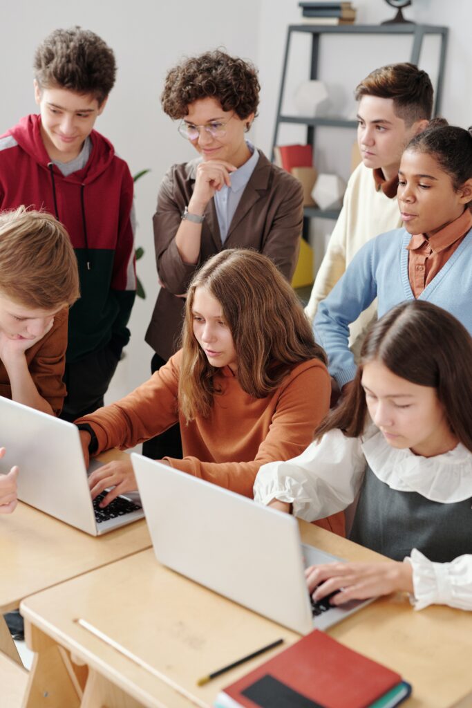 Grupa młodych osób stojąca za dwiema siedzącymi dziewczynkami pracującymi na laptopach.