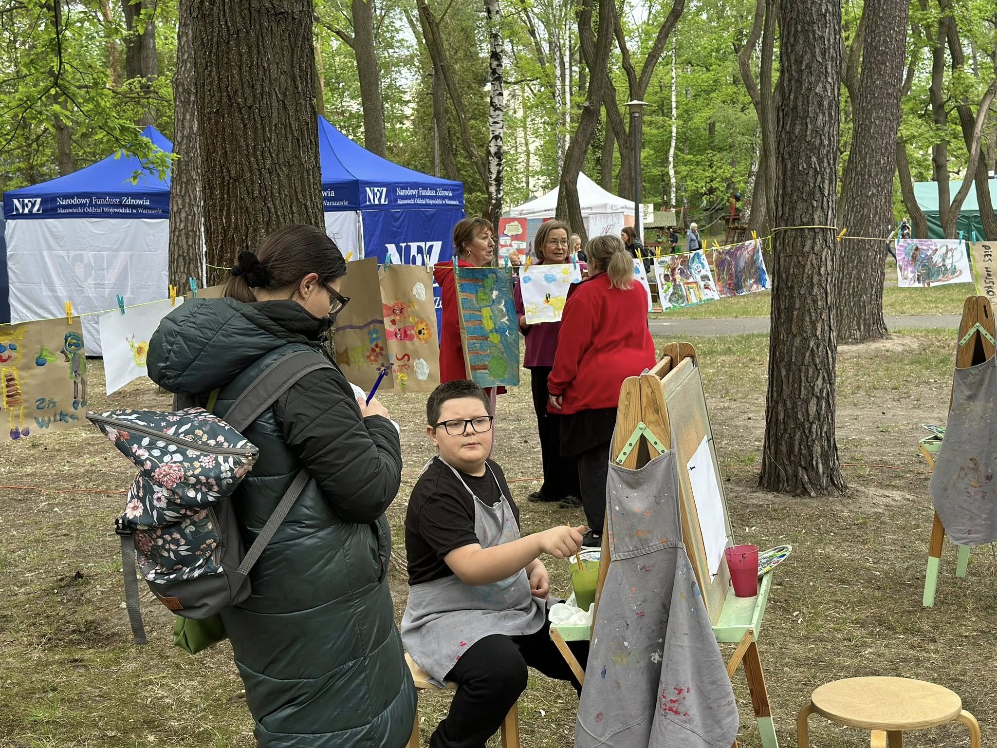 Plenerowe wydarzenie w parku. Na pierwszym planie dziecko maluje obraz przy sztaludze, obok stoi osoba dorosła z plecakiem. Między drzewami rozwieszone są dziecięce prace plastyczne przypięte klamerkami do sznurka. W tle widoczne namioty informacyjne z napisem „NFZ – Narodowy Fundusz Zdrowia, Mazowiecki Oddział Wojewódzki w Warszawie”.