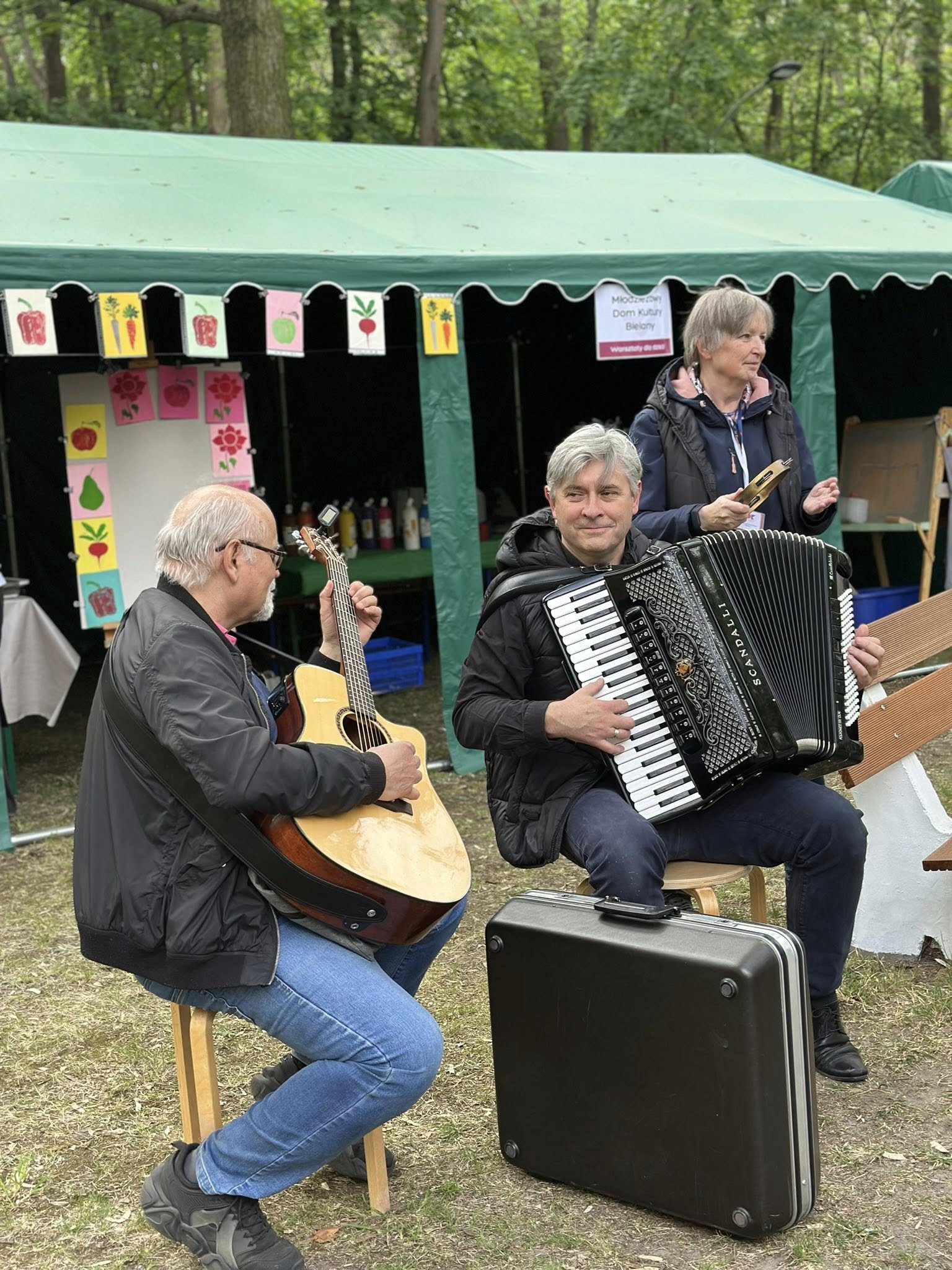 Dwóch muzyków grających w plenerze: jedna osoba na gitarze akustycznej, druga na akordeonie, siedząc na stołkach. W tle zielony namiot warsztatowy z materiałami plastycznymi oraz osoby obserwujące występ.